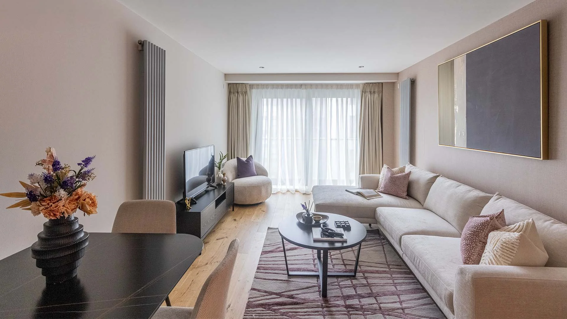 Bright modern living room with neutral sofa, round coffee table, TV unit, and large window with sheer curtains