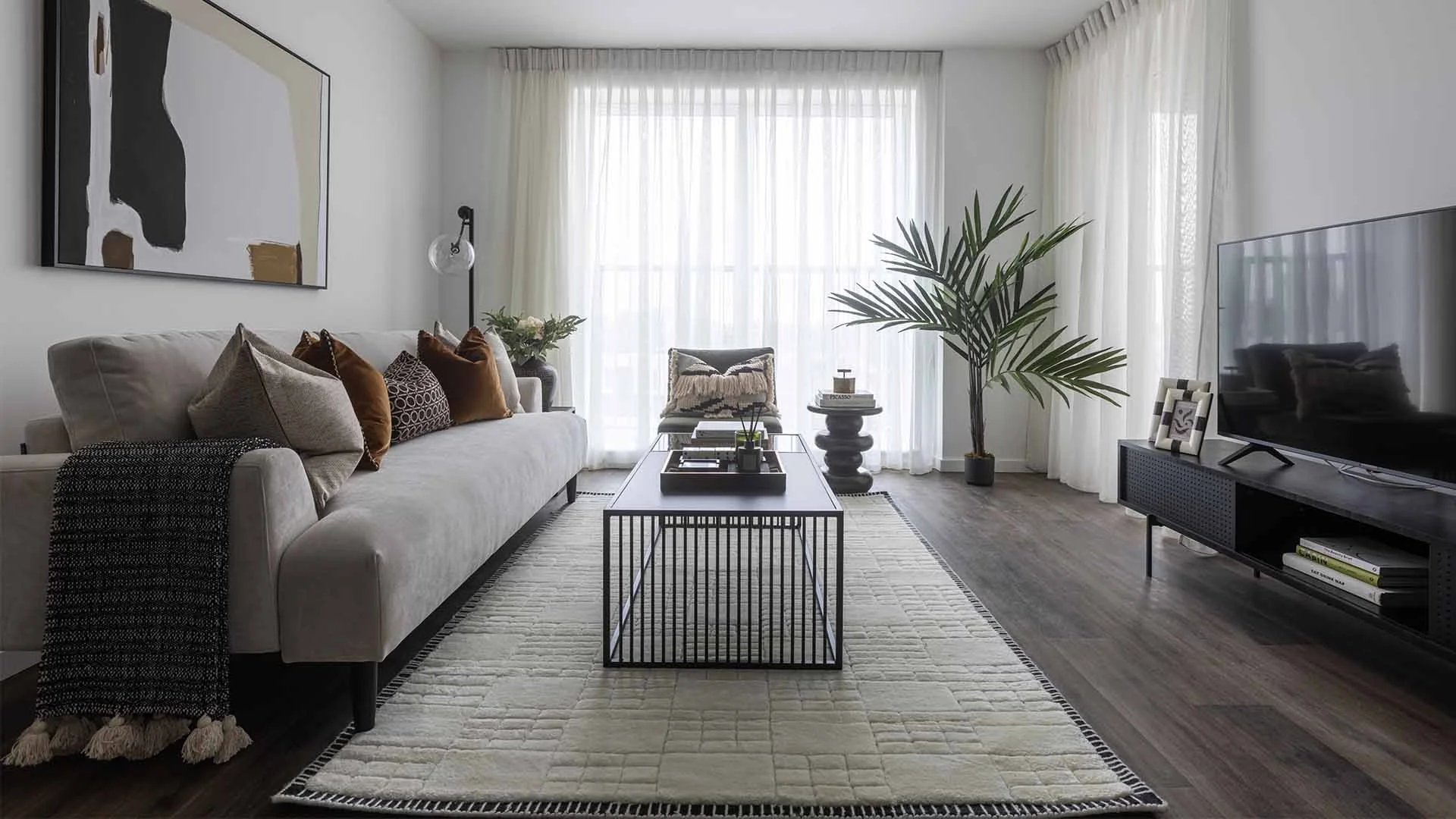 A luxury living room styled by InStyle Direct featuring a light grey sofa with velvet cushions, a black metal slat coffee table, a large indoor palm, and floor-to-ceiling sheer curtains creating a bright, airy space in London.