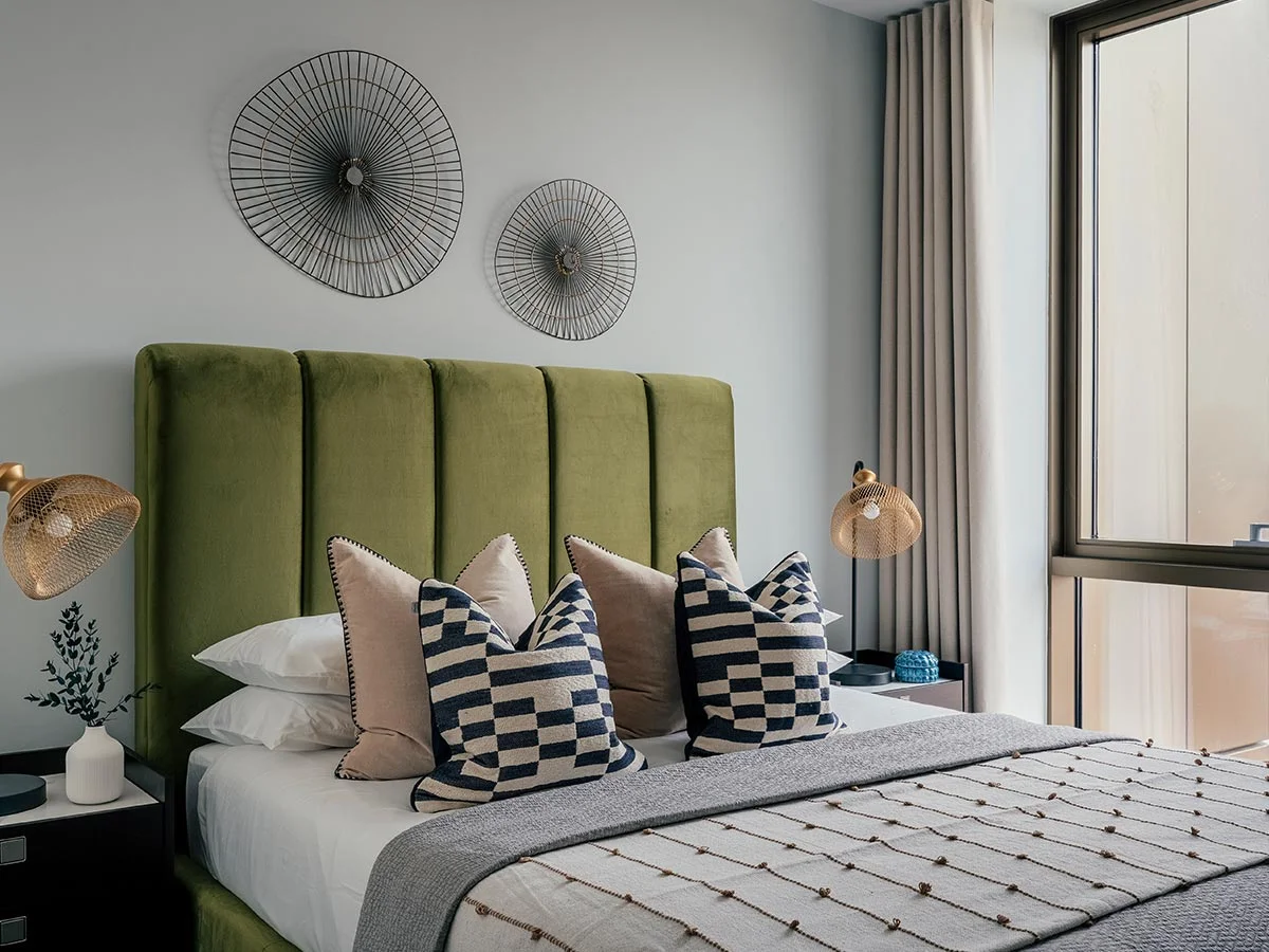 An aspirational show home bedroom designed for property developers, featuring a bold olive green channel-tufted headboard, wire wall art, and gold mesh lamps