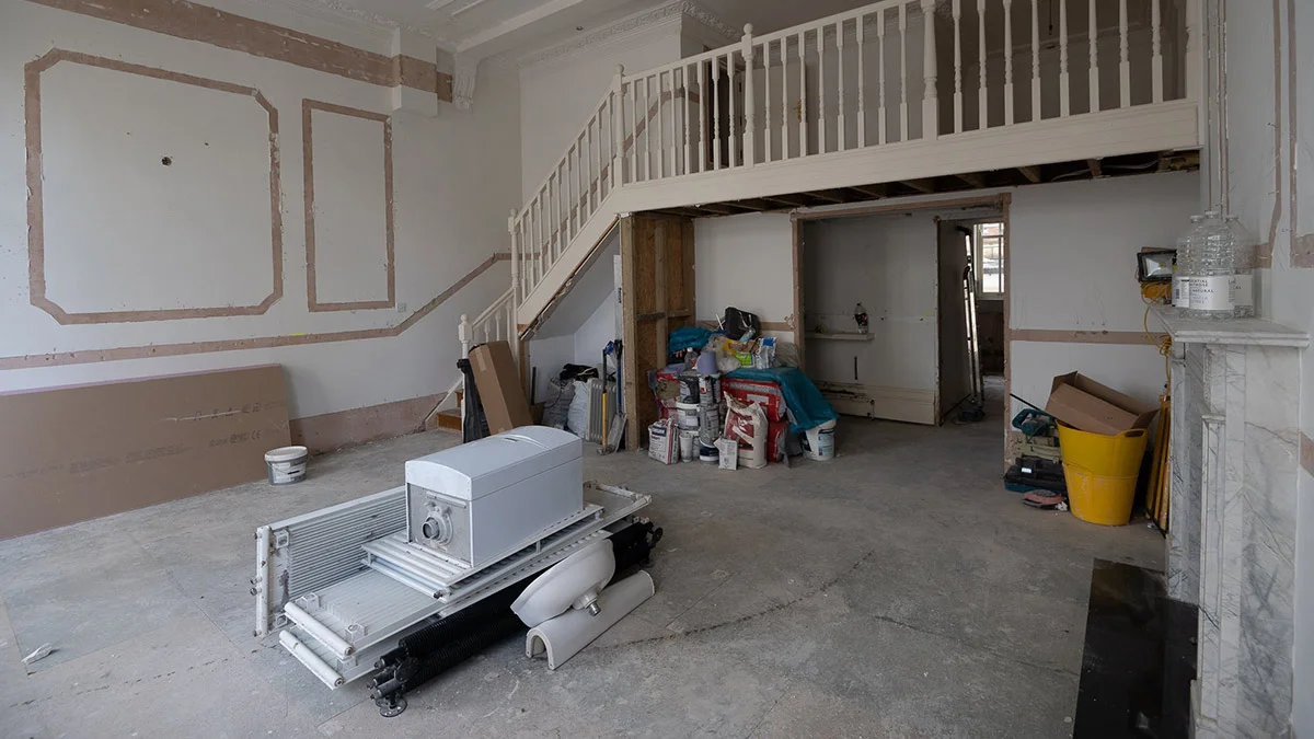 A bare apartment shell in London under major renovation, showing a white staircase leading to a mezzanine, exposed wall moldings, and construction materials scattered across the concrete floor.