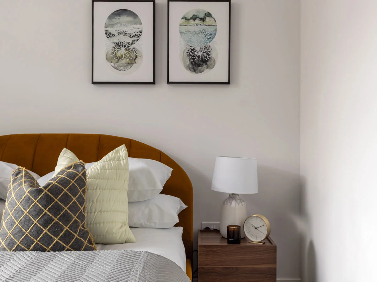 Modern bedroom with upholstered headboard, decorative pillows, and wooden bedside table with lamp and clock.