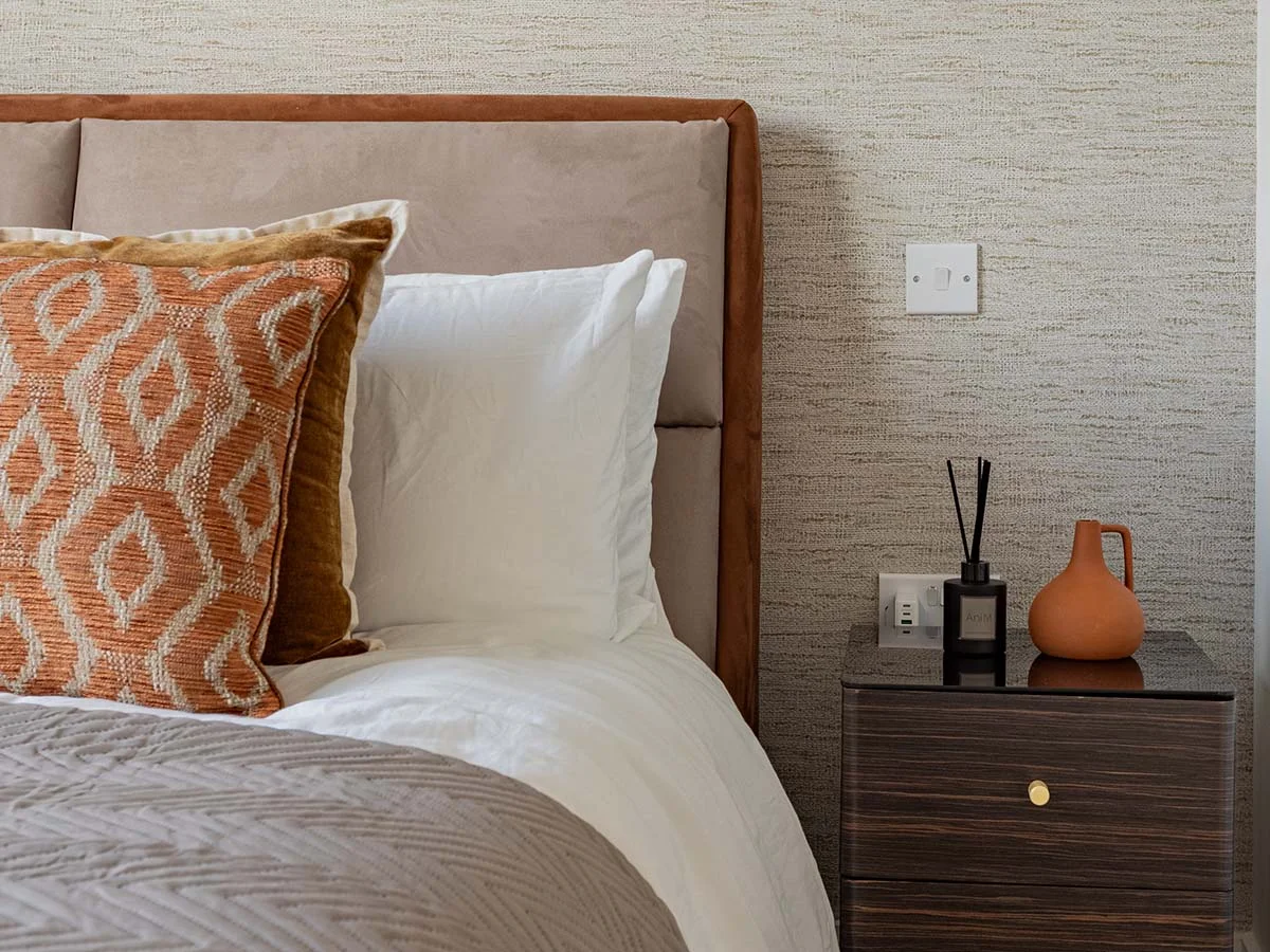 A bespoke bedroom interior by InStyle Direct featuring a taupe upholstered headboard, a dark wood bedside table with a terracotta vase, and a plush bed with an orange geometric patterned cushion in Canary Wharf.
