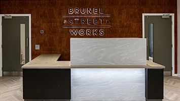 A bespoke commercial reception area styled by InStyle Direct featuring a backlit marble-effect concierge desk against a warm wood-panelled wall with Brunel Street Works branding.