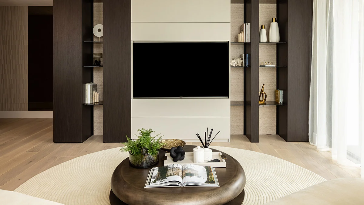 A high-end living area by InStyle Direct featuring a bespoke integrated media wall with dark wood shelving, a large circular bronze coffee table on a cream rug, and minimalist designer decor in Battersea.