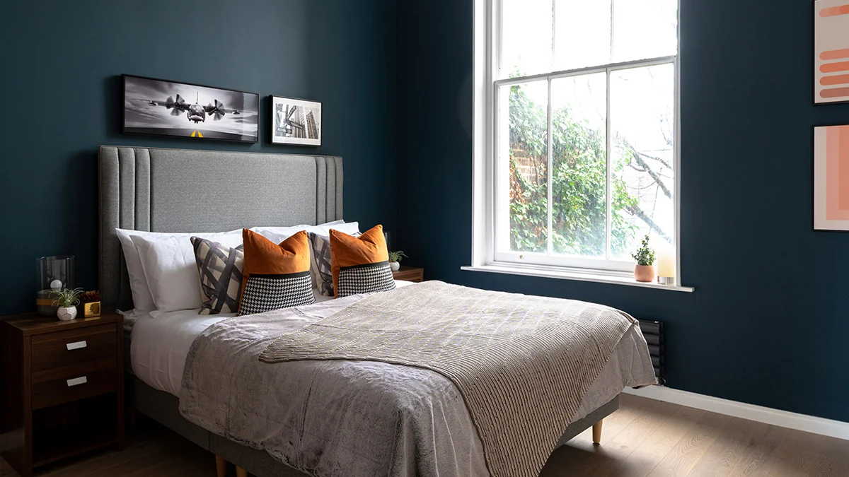 A boutique bedroom styled by InStyle Direct featuring deep navy walls, a tall grey fluted headboard, luxury bedding with orange velvet accent cushions, and aviation-themed wall art in Notting Hill.
