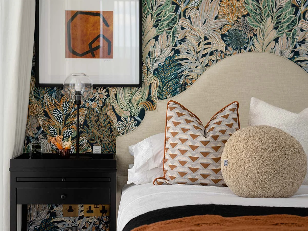 A boutique-style bedroom by InStyle Direct featuring a cream scalloped headboard, a black bedside table with a glass globe lamp, and a vibrant tropical-themed feature wallpaper in London.