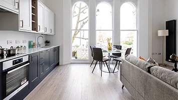 A bright open-plan apartment interior styled by InStyle Direct featuring a sleek navy and white kitchen, a small dining nook with arched windows, and a neutral sofa in Notting Hill.