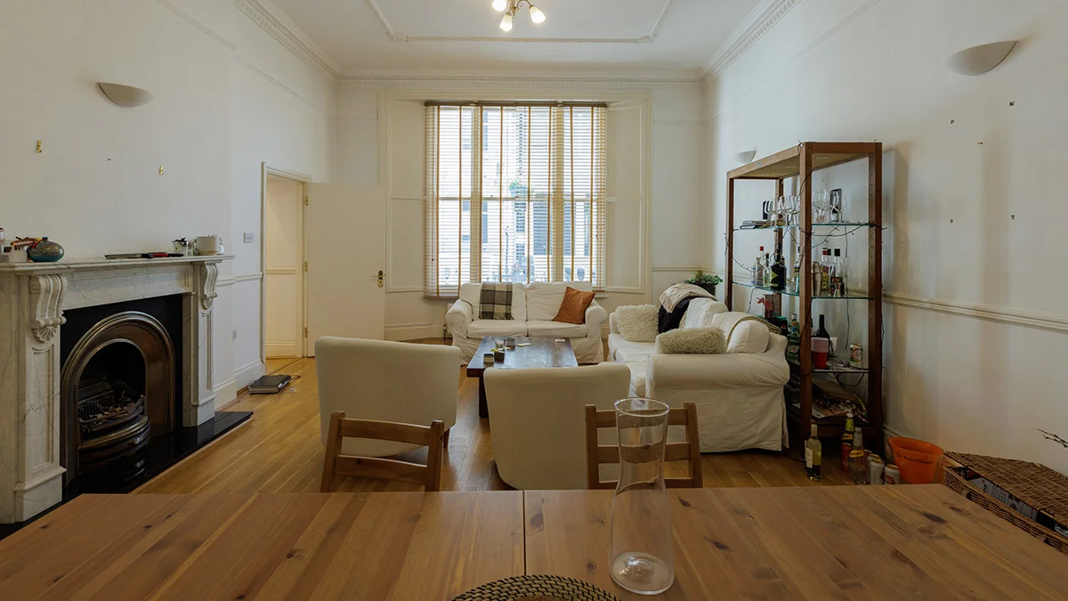 A cluttered and lived-in living room before a luxury redesign, featuring mismatched furniture, basic window blinds, and unorganized shelving in a London apartment.
