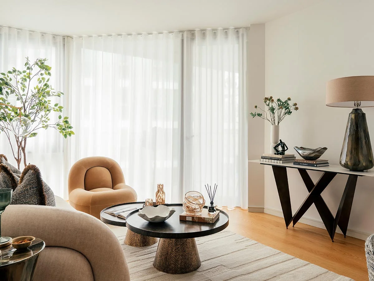 A bright living space featuring a minimalist black metal X-frame console table styled with designer books and a tall green lamp, positioned next to floor-to-ceiling sheer white curtains in a Riverside apartment
