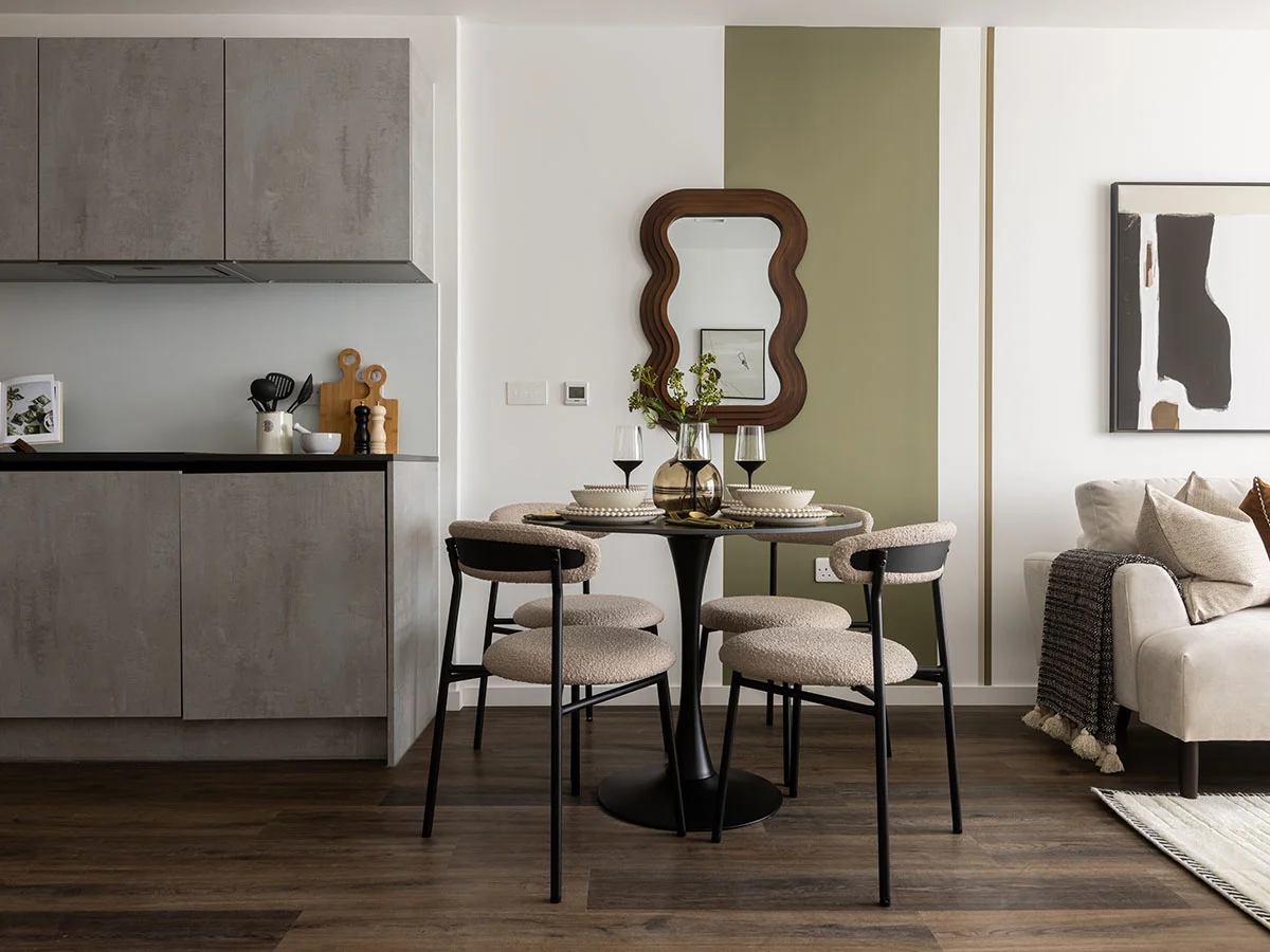 A contemporary dining nook styled by InStyle Direct featuring a round black table with boucle upholstered chairs, an olive green vertical wall stripe with a wavy wooden mirror, and a sleek grey kitchen in Notting Hill.