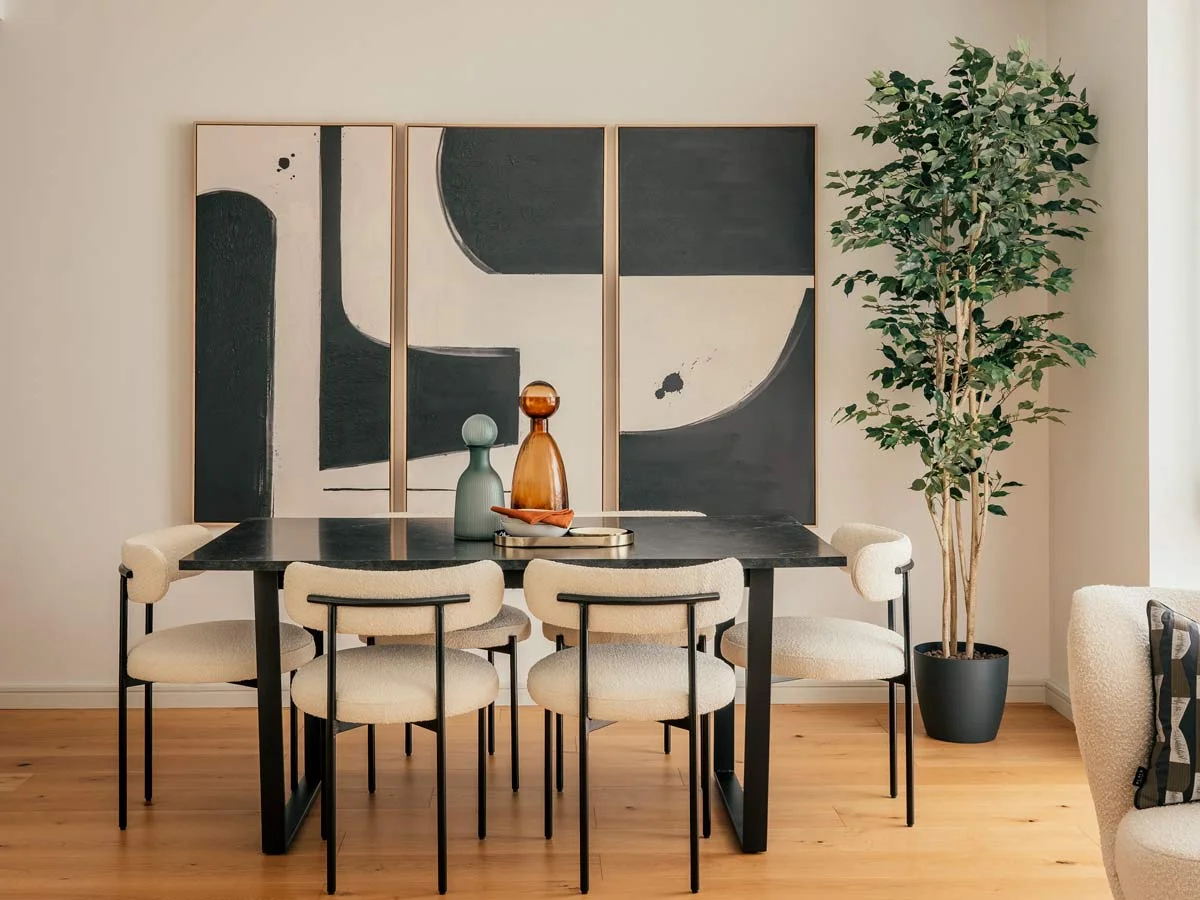Modern dining table with four upholstered chairs, abstract wall art, and potted tree.