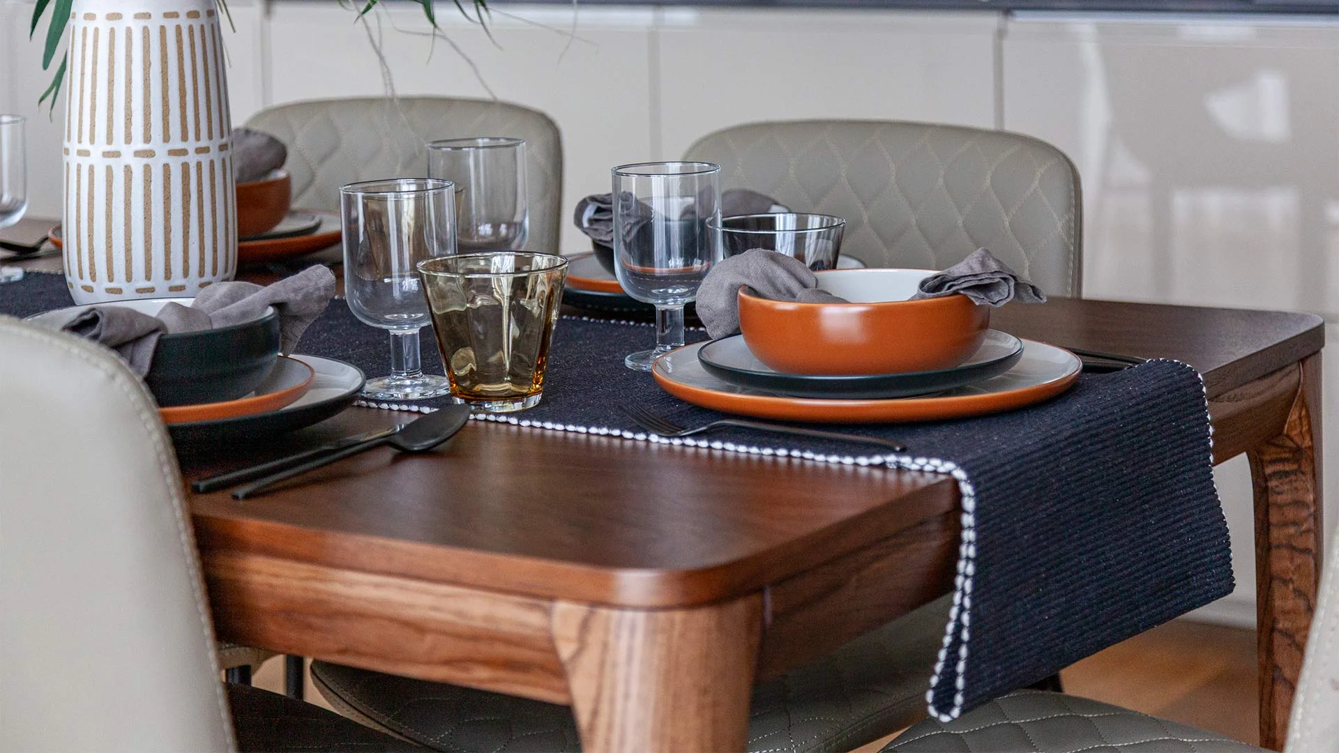 A contemporary dining table set for four by InStyle Direct featuring a dark wood table, quilted grey chairs, terracotta-toned ceramic bowls, and a navy woven table runner in Stratford.