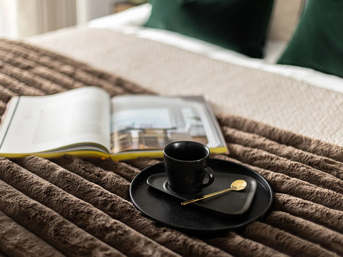 Black coffee cup on tray with gold spoon and open magazine on textured brown bedspread in cozy bedroom setting.