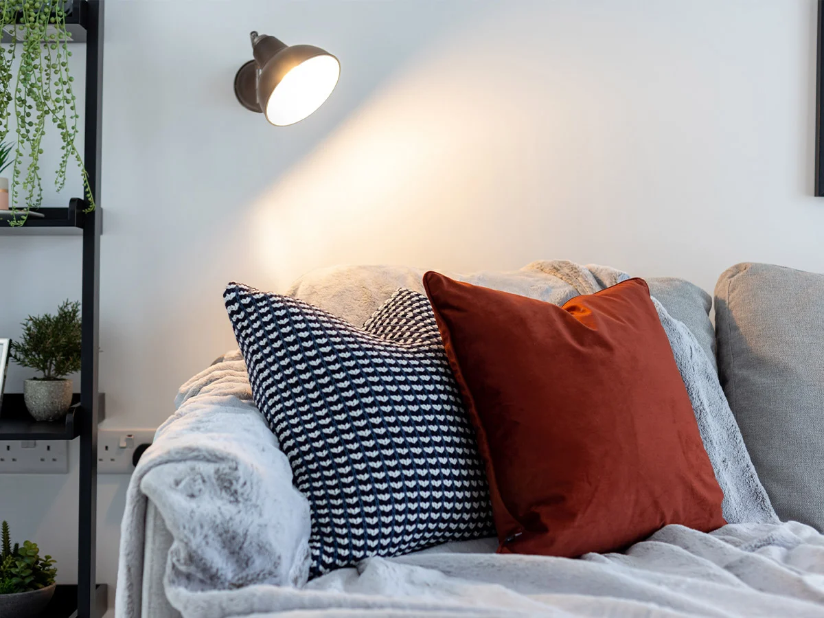 A cozy living room detail by InStyle Direct featuring a grey sofa with a textured navy pillow and a vibrant terracotta velvet cushion, illuminated by a modern industrial wall lamp in Canning Town.