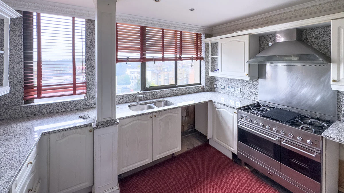 A dated kitchen interior before a luxury makeover, featuring speckled granite countertops and splashbacks, white traditional cabinetry, and red carpeted flooring under bright window light.