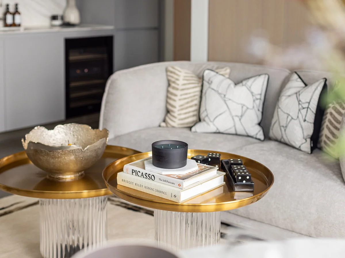 A designer living room space by InStyle Direct featuring a light grey textured sofa with patterned cushions, two tiered circular gold coffee tables displaying art books and a luxury candle, and a sleek modern kitchen in the background in The City of London