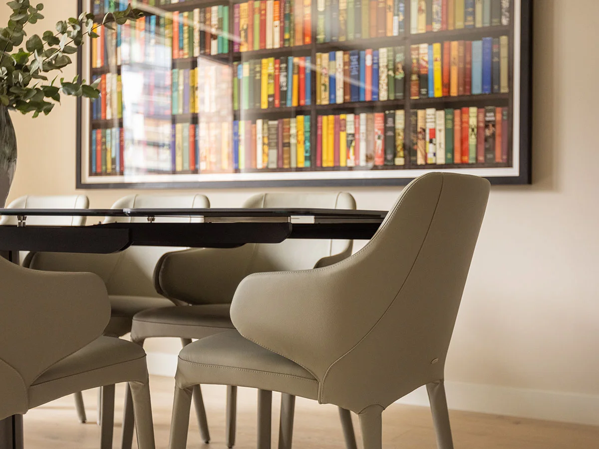 An elegant dining area styled by InStyle Direct featuring sleek grey leather chairs around a glass-top table, with a large framed colorful bookshelf art piece and a fresh eucalyptus arrangement in Chelsea.
