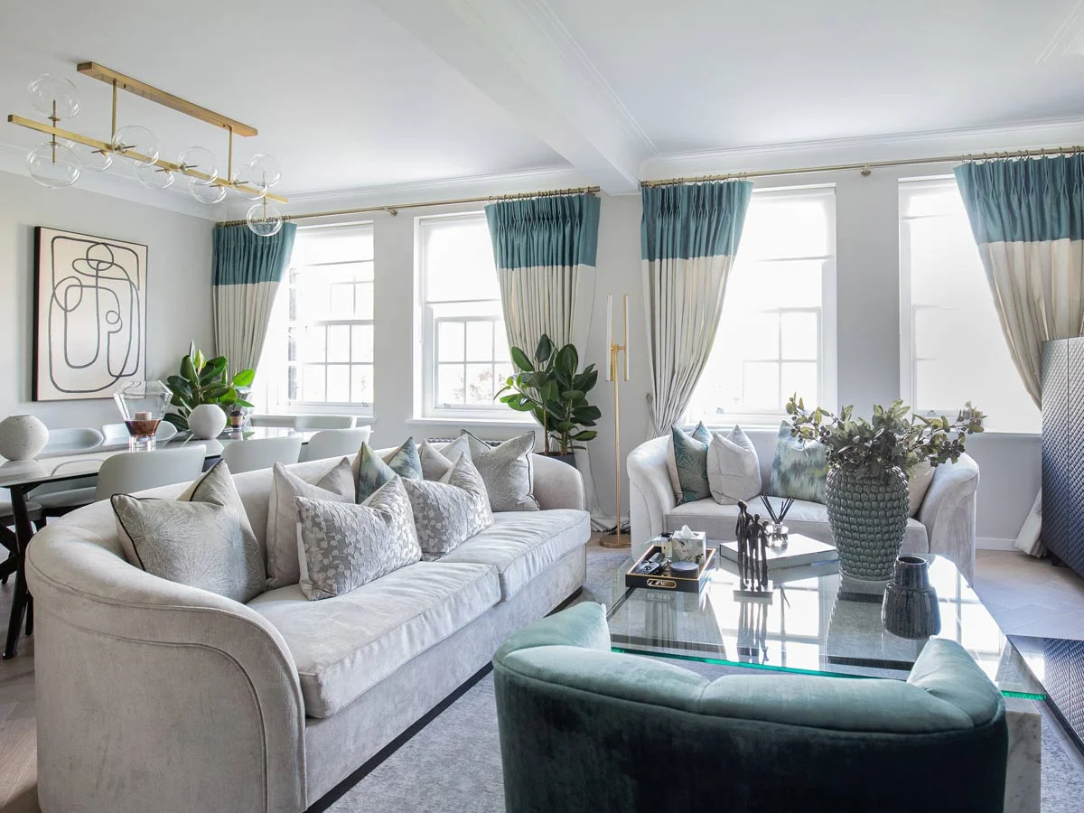 An elegant open-plan living room styled by InStyle Direct featuring a curved grey velvet sofa, teal and cream luxury window dressings, a glass-top coffee table, and a modern gold chandelier in Hampstead.