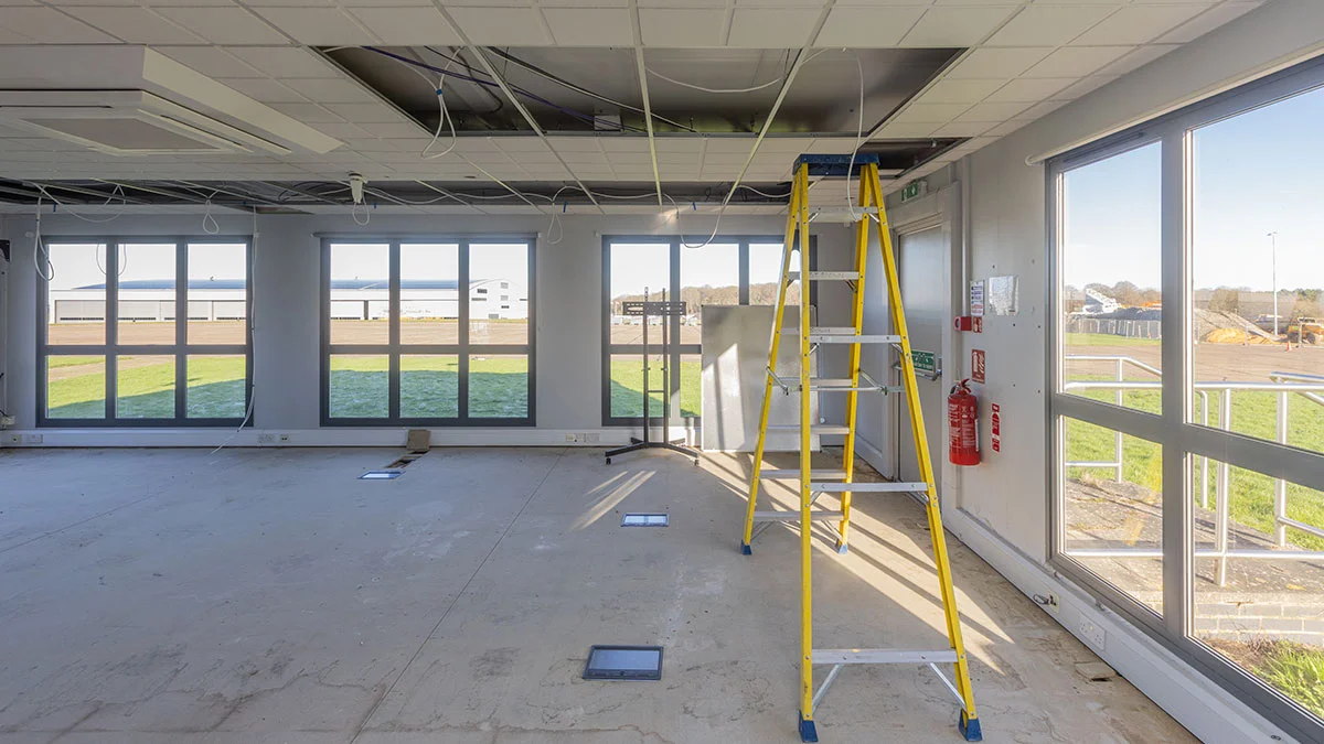 An empty commercial office shell under construction, showing bare concrete floors, exposed ceiling wiring, and a yellow ladder before a professional furniture installation by InStyle Direct.