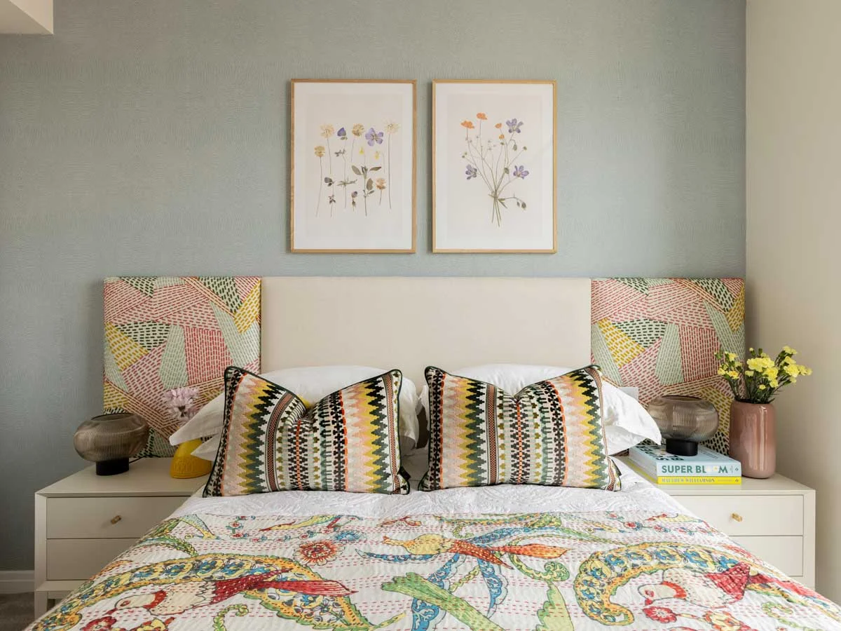 A master bedroom in the Horlicks Quarter, Slough, featuring an amber velvet scalloped headboard, geometric patterned cushions, a dark wood nightstand with a marble lamp, and abstract botanical artwork