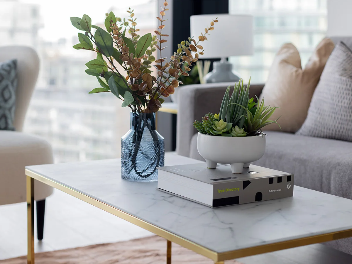 Close-up of a marble and gold coffee table styled for an investor furniture package, featuring a blue glass vase with greenery, a white potted succulent, and designer interior books in a London apartment.