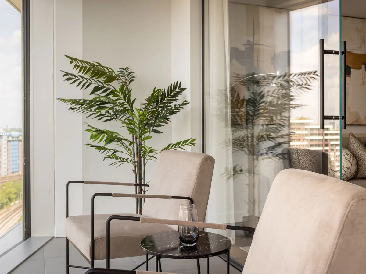 Modern balcony seating area with beige lounge chairs, black metal frame, side table, and indoor plant beside glass doors in a contemporary apartment