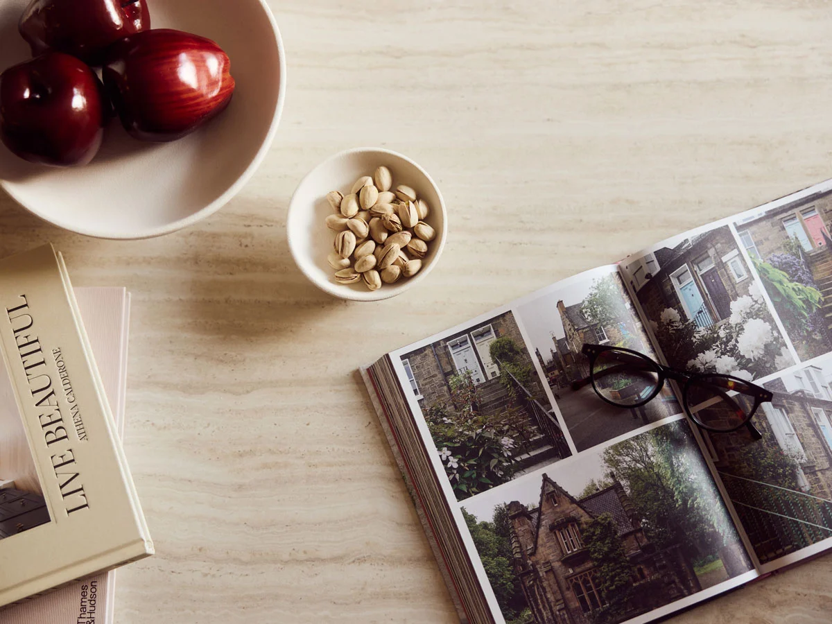 Open book with house photos, glasses, apples, pistachios, and stacked books on a table.