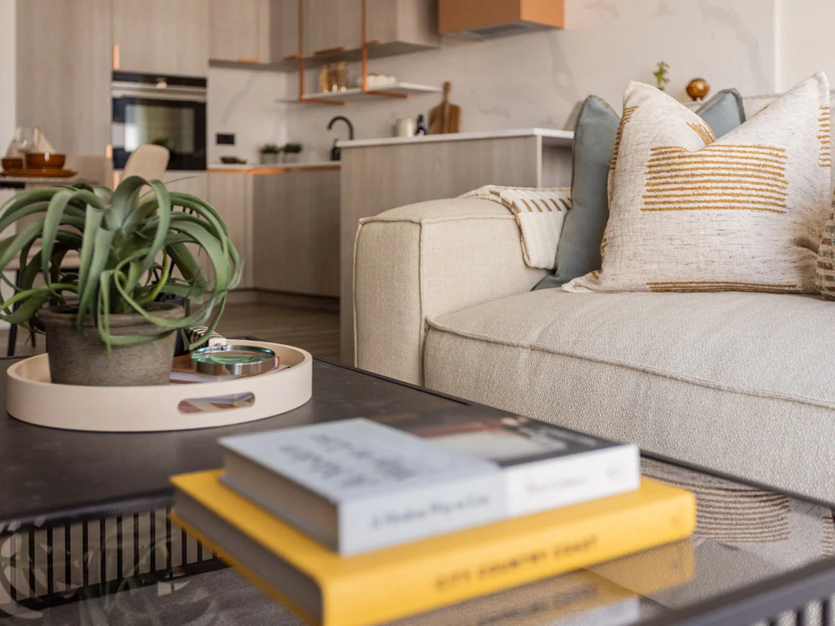 Modern living room with beige sofa, decorative pillows, coffee table with books, and open kitchen in background.