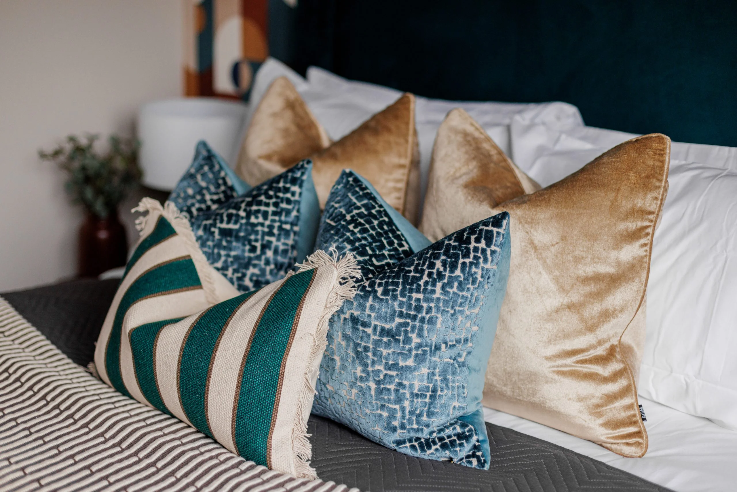 A luxury bedding arrangement styled by InStyle Direct featuring a layered selection of plush gold velvet cushions, textured teal pillows, and a striped accent cushion on a contemporary grey bed in Greenwich.