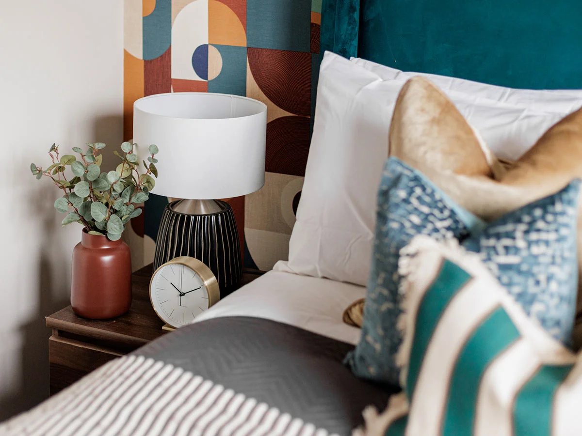 A luxury bedroom corner styled by InStyle Direct featuring a plush teal velvet headboard, a dark wood bedside table with a modern black and white lamp, and retro geometric patterned wallpaper in shades of orange, blue, and brown in Islington