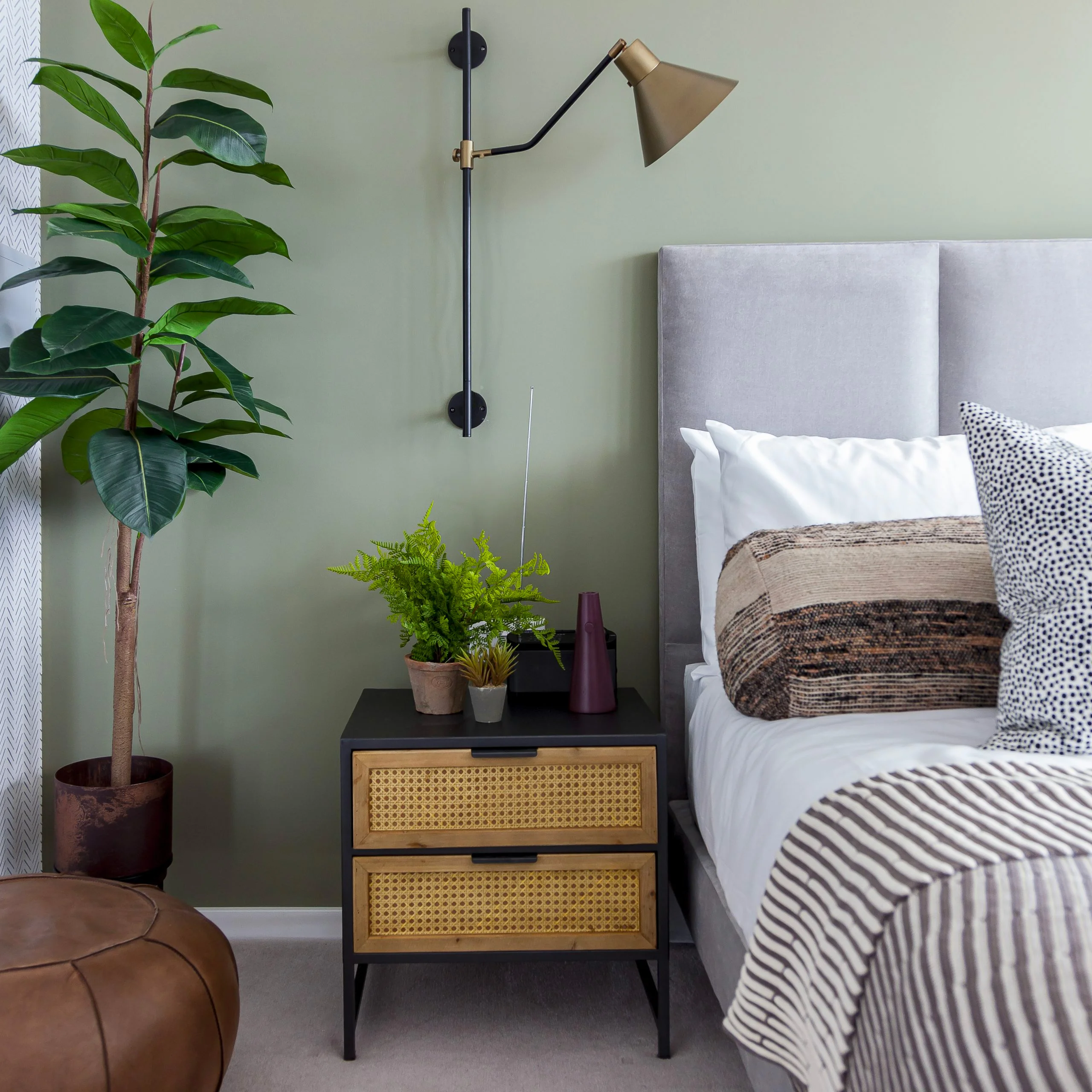A luxury bedroom styled by InStyle Direct featuring a sage green feature wall, a light grey upholstered headboard, a black bedside table with rattan drawers, and a large indoor rubber plant in Marylebone.
