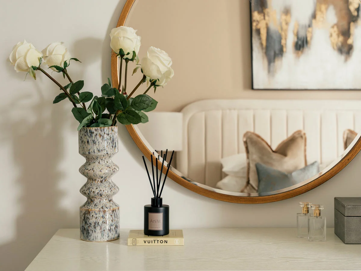 White roses in textured ceramic vase on console table with round mirror, reed diffuser, and bedroom reflection in background.