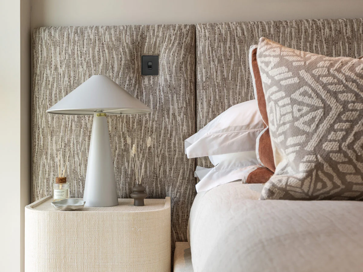 A luxury bedroom styled by InStyle Direct featuring a floor-to-ceiling textured grey fabric headboard, a sleek conical grey bedside lamp on a curved neutral nightstand, and plush layered bedding with geometric patterned cushions in Kensington