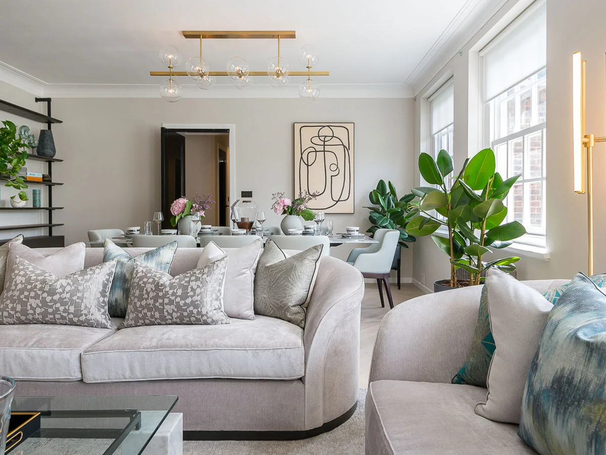 A luxury open-plan lounge styled by InStyle Direct featuring a curved grey velvet sofa with patterned cushions, a modern gold chandelier, and a designer dining area with a large abstract portrait in Hampstead.