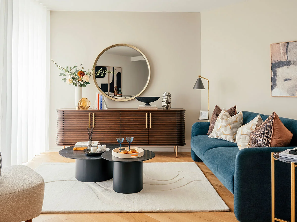 A sophisticated living area in Battersea featuring a deep blue velvet sofa, a slatted dark wood sideboard with a large circular gold mirror, and twin marble-top coffee tables styled by InStyle Direct
