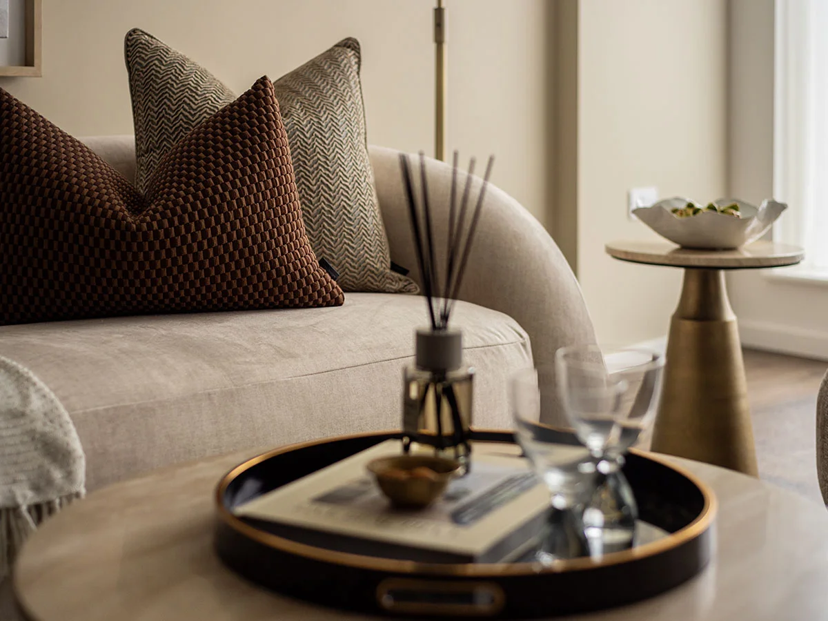 A luxury living room detail styled by InStyle Direct featuring a curved beige sofa with textured brown and patterned cushions, a reed diffuser on a marble coffee table, and a gold pedestal side table in Notting Hill.