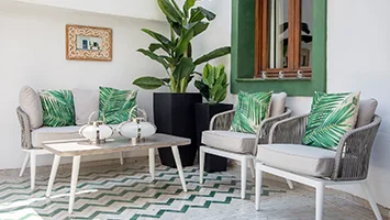 A Mediterranean-style outdoor patio styled by InStyle Direct featuring light grey rope-effect armchairs with vibrant tropical palm leaf cushions, a wooden coffee table, and large potted banana plants.
