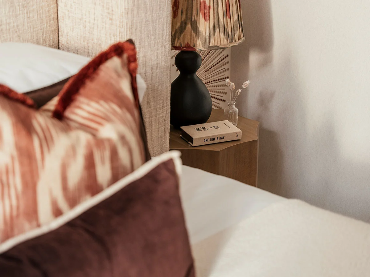 A minimalist bedside corner styled by InStyle Direct featuring a wooden hexagonal nightstand, a black designer lamp with an ikat pattern shade, and a small glass vase with dried botanicals