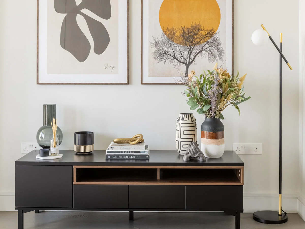 A minimalist black media console styled by InStyle Direct featuring a collection of designer vases, art books, and a gold decorative object, positioned beneath two large framed abstract prints with a modern black and gold floor lamp in Mayfair