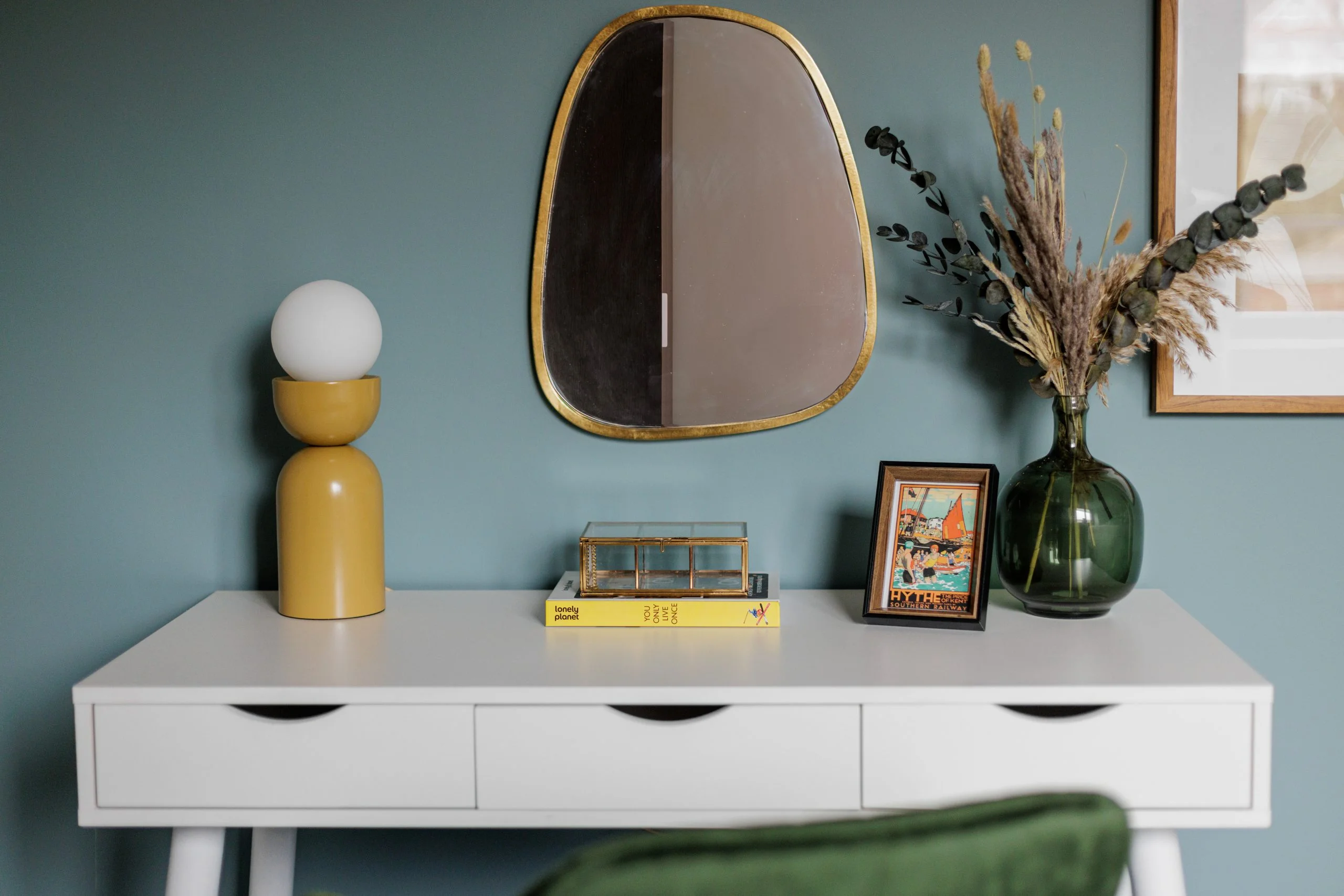 A minimalist home office desk styled by InStyle Direct featuring a sleek white workspace, a modern mustard-yellow table lamp, an asymmetric gold-framed mirror, and a green glass vase with dried botanicals in Chelsea.