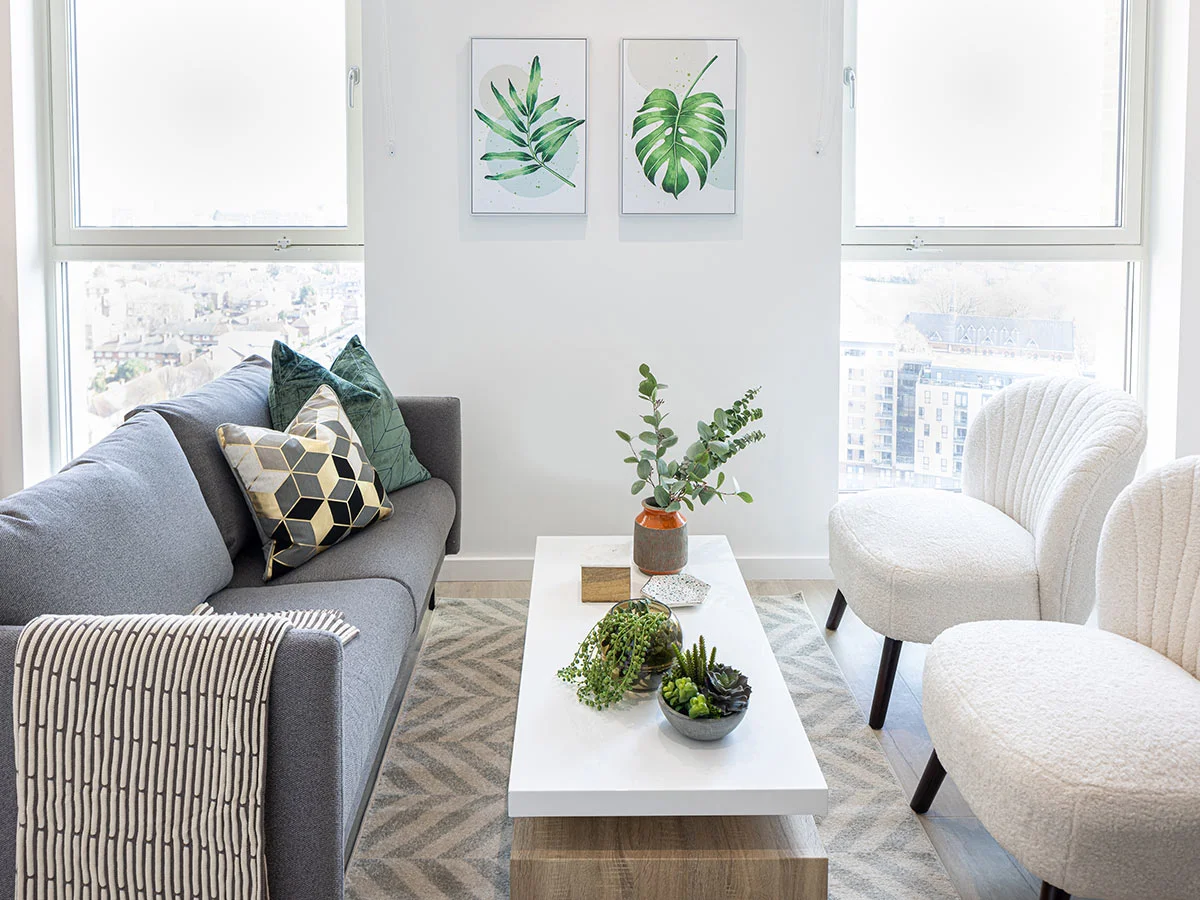 A bright modern living room styled by InStyle Direct featuring a grey sofa with geometric cushions, two white bouclé armchairs, a marble coffee table with succulents, and botanical leaf wall art.