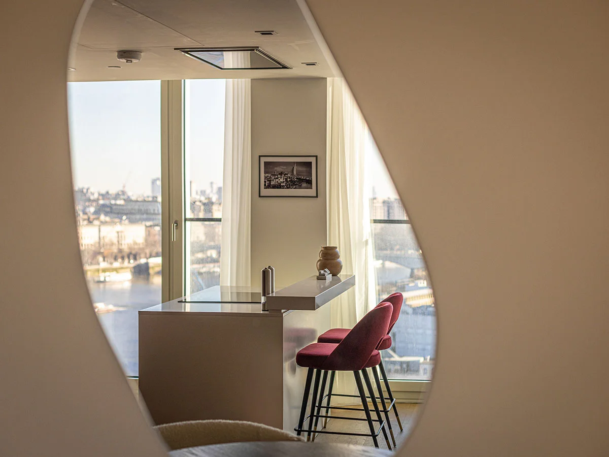 A modern minimalist kitchen breakfast bar styled by InStyle Direct featuring deep red velvet bar stools and a sleek white countertop, viewed through a circular mirror with the River Thames in the background at Southbank Tower.