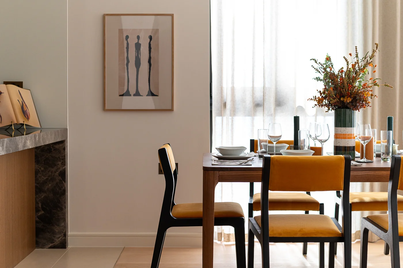 A modern dining space by InStyle Direct featuring a wooden table with mustard yellow upholstered chairs, a tall vase of autumnal flowers, and a large minimalist art piece in Holborn.