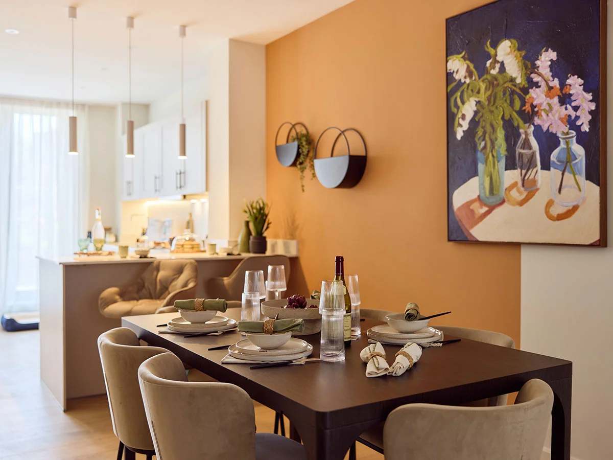 A modern dining space at New Fountainbridge featuring a dark wood table, beige velvet chairs, and a vibrant orange feature wall with floral artwork.
