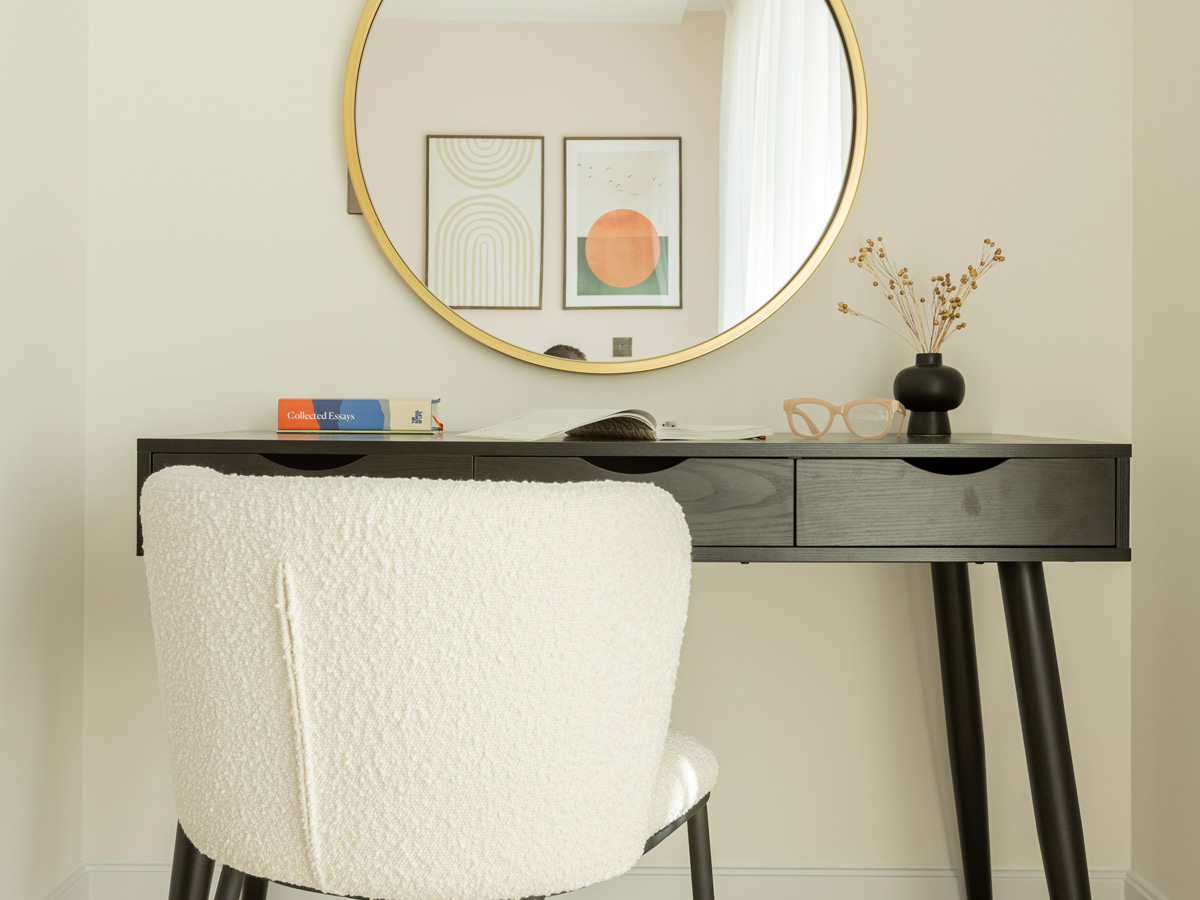 A modern home office nook styled by InStyle Direct featuring a sleek black wooden desk, a white textured boucle chair, and a large gold-framed circular mirror reflecting minimalist wall art.