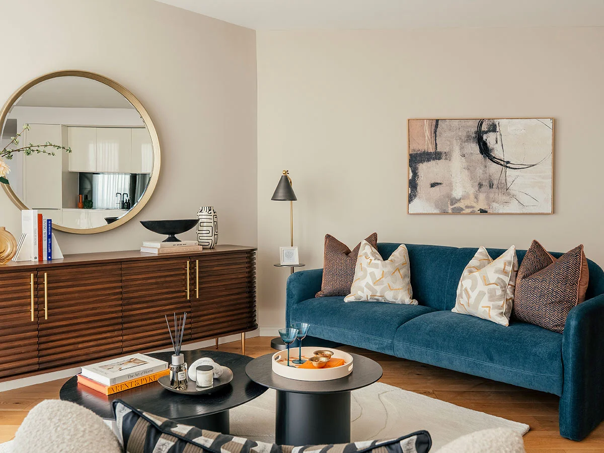 A contemporary living room at Riverside featuring a blue velvet sofa, a slatted dark wood console, and a large round mirror