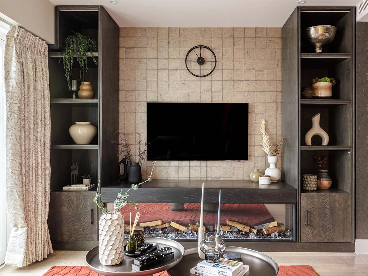 A modern media wall styled by InStyle Direct featuring a flat-screen TV mounted on a grid-patterned wallpaper, bespoke dark wood shelving with curated decor, and a contemporary linear fireplace in The Strand.