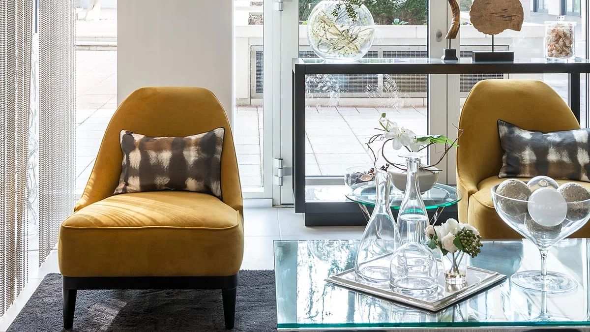 A modern living space styled by InStyle Direct featuring vibrant mustard yellow velvet accent chairs with patterned cushions, a glass-top coffee table with designer glassware, and large floor-to-ceiling windows at Imperial Wharf.