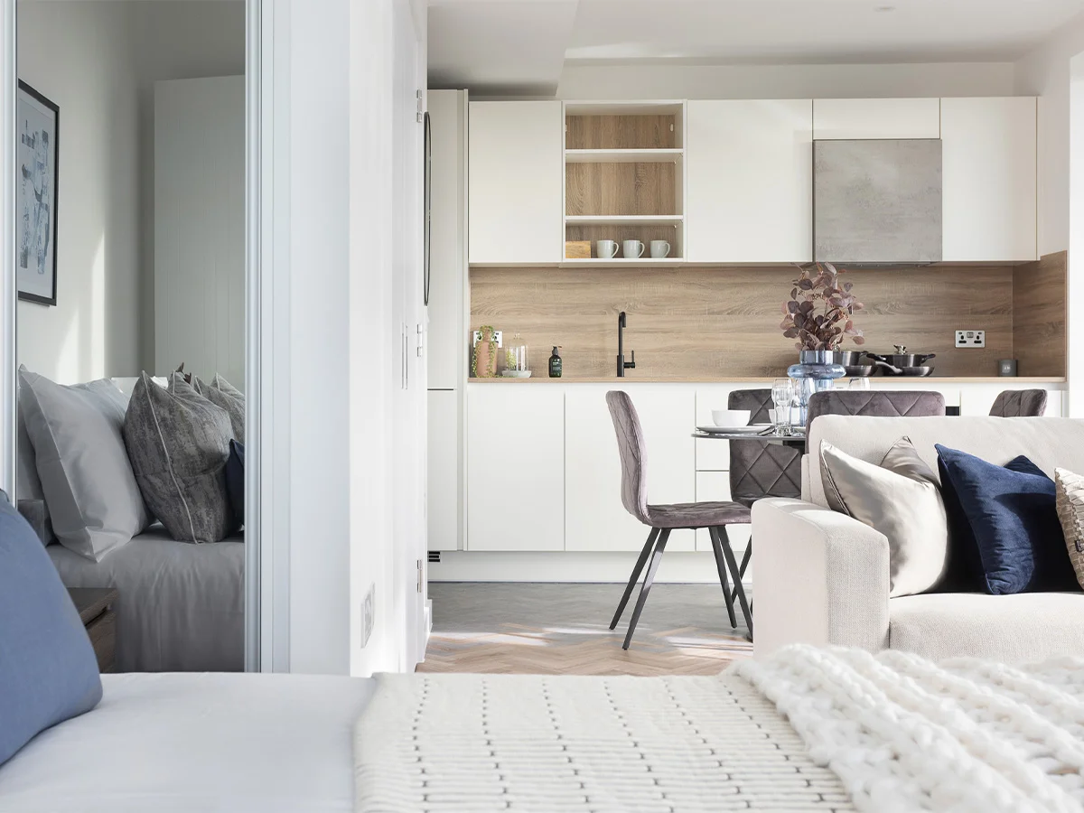 A bright open-plan apartment interior styled by InStyle Direct featuring a modern white kitchen with wood accents, a neutral living area with navy velvet cushions, and a cozy bedroom view in Bow.