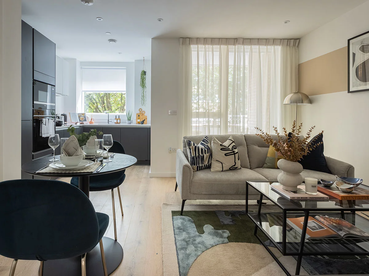 A small open-plan apartment featuring a light grey sofa, a dark navy kitchen in the background, and a colourful modern rug defining the living area